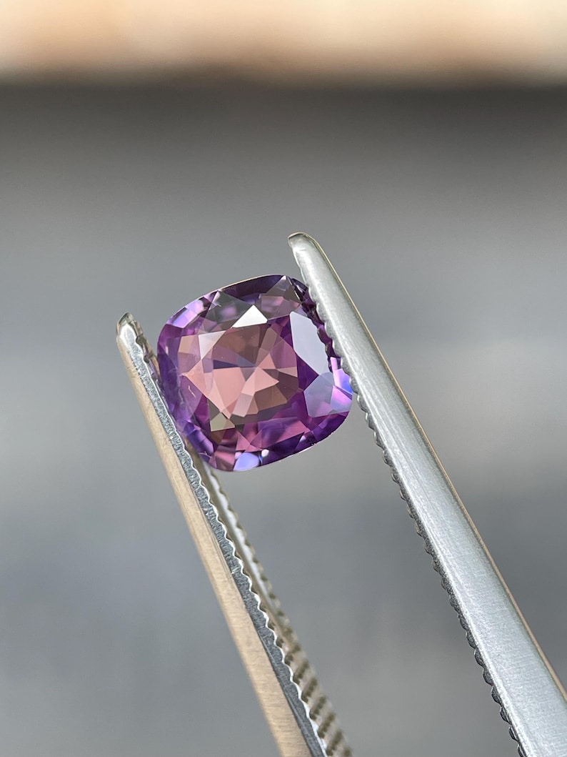 May include: A cushion-cut gemstone with a blend of purple and pink hues is held by a pair of silver tweezers. The gem's facets reflect light, creating a sparkling effect. The background is a soft, blurred gray.