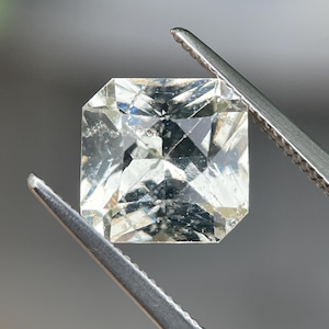 May include: A square-cut, transparent gemstone with a faceted surface that reflects light. The stone features internal inclusions and a clear, colourless appearance, held by metal tweezers that grip the top and bottom edges of the gem.