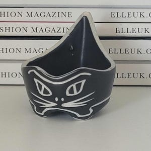 May include: A black ceramic cat-shaped dish with white facial details. The dish has a triangular form with a curved edge. The cat's face is shown with white eyes, nose, and whiskers. The dish is likely for holding small items.