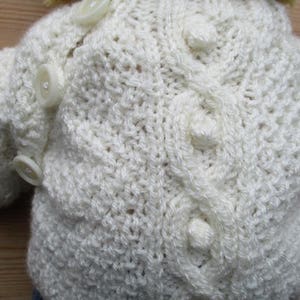 May include: White knitted cardigan with a cable knit pattern and white buttons.