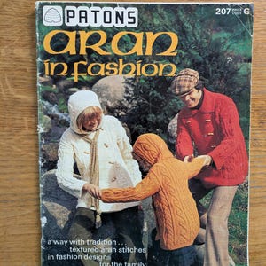 以下が含まれることがあります： Patons Aran in Fashionのヴィンテージ編み物パターンブックレット。表紙には、白、オレンジ、赤のケーブル編みのフード付きセーターやジャケットを着用し、トグルボタンで留めた3人が自然の屋外を背景に写っている。