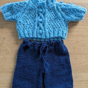 May include: A knitted baby outfit in blue and navy. The top is a light blue cardigan with a cable knit pattern and buttons. The bottom is a pair of navy blue shorts.
