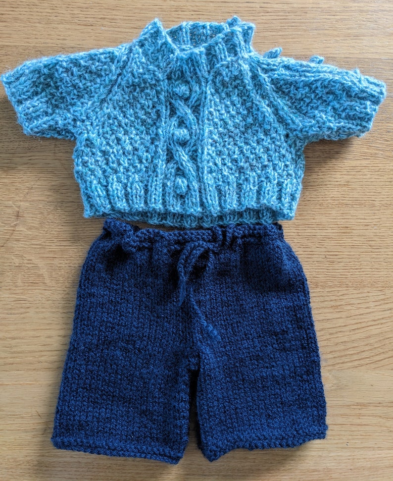 May include: A knitted baby outfit in blue and navy. The top is a light blue cardigan with a cable knit pattern and buttons. The bottom is a pair of navy blue shorts.