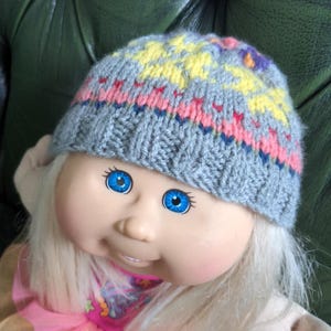 May include: A grey knitted hat with a colourful geometric pattern. The hat is worn by a doll with blonde hair and blue eyes.