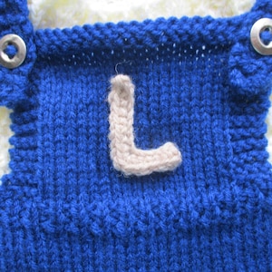May include: Blue knitted baby overalls with a white knitted trim and a beige knitted letter L on the front.