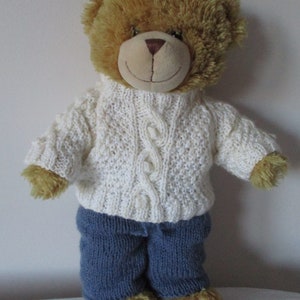 May include: A brown teddy bear wearing a white knitted sweater with a cable knit pattern and blue knitted trousers.