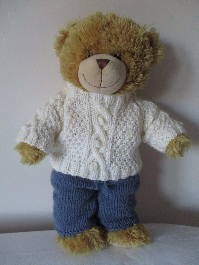 May include: A brown teddy bear wearing a white knitted sweater with a cable knit pattern and blue knitted trousers.