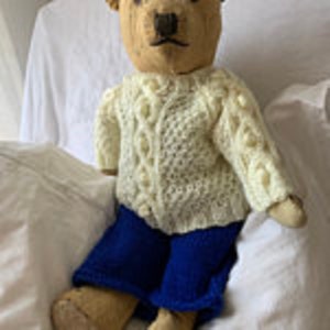 May include: A vintage teddy bear wearing a white knitted sweater with a cable knit pattern and blue trousers.