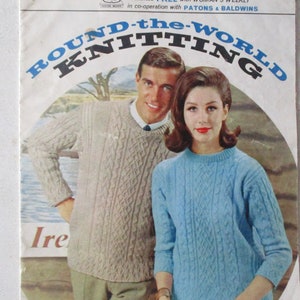 以下が含まれることがあります： ケーブル編みのセーターを着たカップルが表紙に描かれた、ビンテージの編み物パターンブックレット。タイトルは「Round-the-World Knitting」で、テキストには「Presented FREE with WOMAN'S WEEKLY in co-operation with PATONS & BALDWINS」と記載されています。