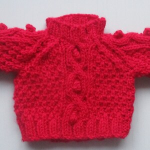 May include: A red knitted baby sweater with a cable knit pattern and bobbles on the shoulders.