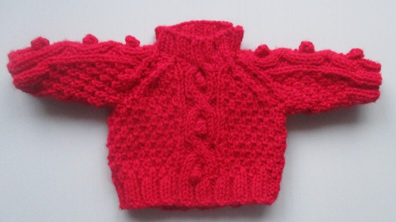 May include: A red knitted baby sweater with a cable knit pattern and bobbles on the shoulders.