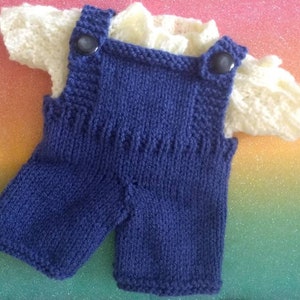 May include: A knitted baby outfit with a blue overall and a white shirt. The overall has two buttons on the straps.
