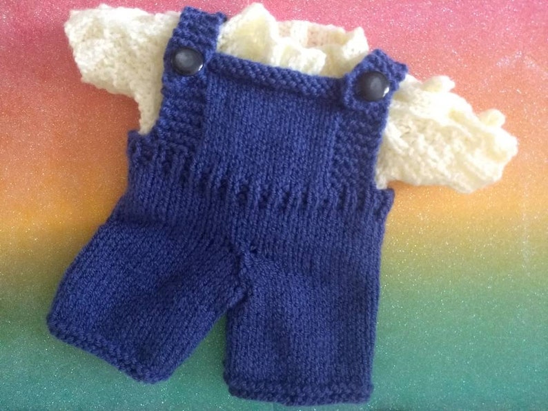 May include: A knitted baby outfit with a blue overall and a white shirt. The overall has two buttons on the straps.