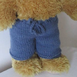 May include: A brown teddy bear wearing blue knitted trousers with a drawstring waist.