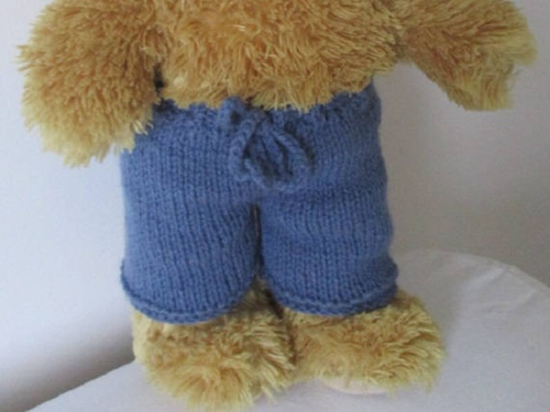 May include: A brown teddy bear wearing blue knitted trousers with a drawstring waist.