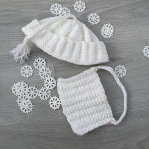 May include: A white knitted baby hat with a tassel and a matching face covering. The set is surrounded by white snowflake decorations. The hat and face covering are made of a soft, textured yarn.