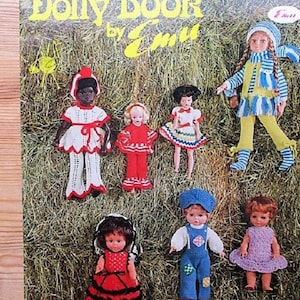 May include: A book titled "Dolly Book by Emi" with a cover featuring a variety of knitted dolls in different outfits. The dolls are standing on a background of hay.