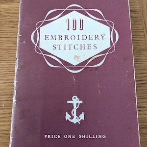 May include: A maroon softcover book titled 100 Embroidery Stitches with white text and a white anchor graphic centred on the front cover. The cover features a decorative white geometric frame around the title and the text Price One Shilling.