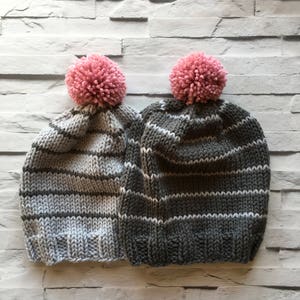 Puede incluir: Dos gorros de punto con rayas horizontales y un pomp&oacute;n rosa en la parte superior. Un gorro es gris claro con rayas gris oscuro, mientras que el otro es gris carb&oacute;n oscuro con rayas gris claro. Ambos tienen un borde acanalado en la base.
