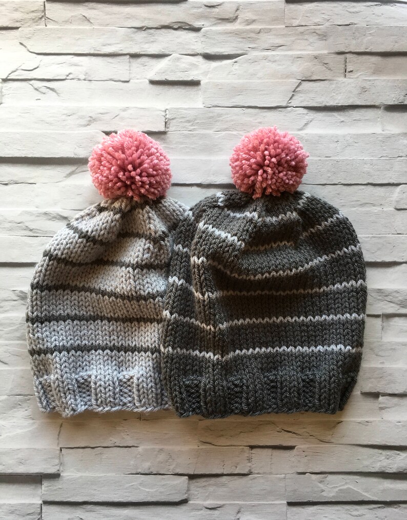 Puede incluir: Dos gorros de punto con rayas horizontales y un pomp&oacute;n rosa en la parte superior. Un gorro es gris claro con rayas gris oscuro, mientras que el otro es gris carb&oacute;n oscuro con rayas gris claro. Ambos tienen un borde acanalado en la base.