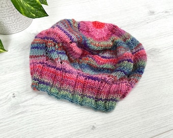 Könnte beinhalten: Eine bunte, handgestrickte Beanie-Mütze. Die Mütze hat eine lebendige Mischung aus rosa, roten, lila, blauen und grünen Garnen. Die Mütze ist aus weichem, strukturiertem Garn gefertigt und ist zum Tragen auf dem Kopf bestimmt.