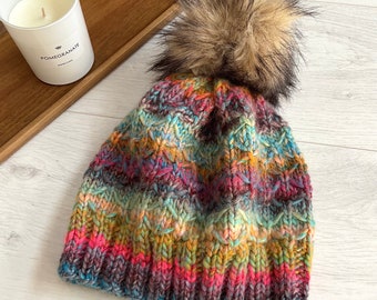 Könnte beinhalten: Eine mehrfarbige Strickmütze mit einem Kunstfell-Bommel. Die Mütze besteht aus blauen, rosa, gelben und braunen Garnen. Der Bommel ist braun und beige. Ein Winteraccessoire.