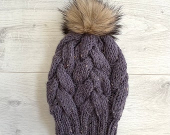 Könnte beinhalten: Eine gestrickte lila Mütze mit Zopfmuster und einem Kunstfell-Bommel oben. Die Mütze ist aus gesprenkeltem Garn gefertigt und für kaltes Wetter konzipiert. Der Bommel hat eine hellbraune Farbe.