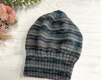 Könnte beinhalten: Eine gestrickte Beanie-Mütze mit horizontalen Streifen in Grau-, Braun-, Türkis- und Blautönen. Die Mütze ist aus weichem Garn gefertigt und hat einen umgeschlagenen Saum. Die Mütze ist ein Winteraccessoire.