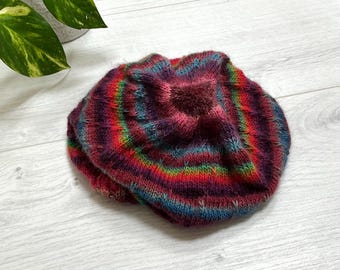 Könnte beinhalten: Eine runde, gestrickte Baskenmütze mit einem lebendigen, mehrfarbigen Verlaufsmuster. Das Garn zeigt horizontale Streifen in Dunkelviolett, leuchtendem Rot, Orange und Blaugrün, was eine weiche, strukturierte Optik mit einem gerafften Mittelpunkt erzeugt.
