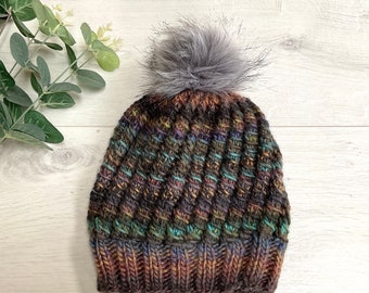 Könnte beinhalten: Eine gestrickte Beanie-Mütze mit einem strukturierten, diagonalen Muster in Braun-, Grün- und Lilatönen. Die Mütze ist mit einem flauschigen, grauen Kunstfell-Bommel versehen. Ein Winteraccessoire.