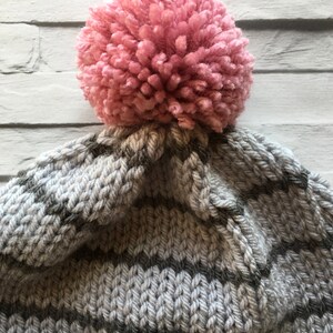 Puede incluir: Un gorro de punto con rayas horizontales en tonos alternos de gris claro y gris oscuro. La parte superior del gorro est&aacute; rematada con un pomp&oacute;n grande, redondo y texturizado en un suave color rosa empolvado, sujeto a la corona.