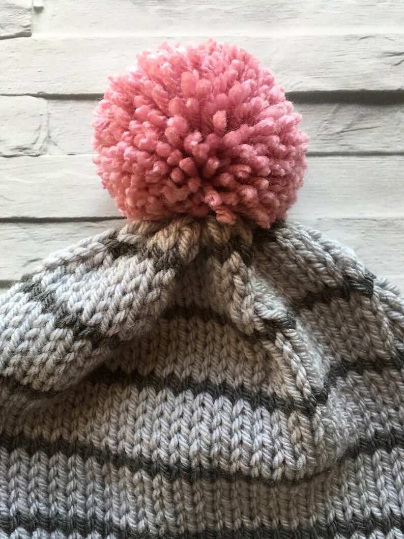 Puede incluir: Un gorro de punto con rayas horizontales en tonos alternos de gris claro y gris oscuro. La parte superior del gorro est&aacute; rematada con un pomp&oacute;n grande, redondo y texturizado en un suave color rosa empolvado, sujeto a la corona.