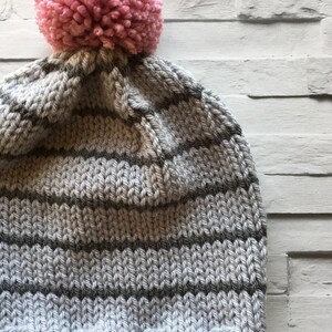 Puede incluir: Un gorro de punto con un borde acanalado y rayas horizontales en tonos alternos de gris claro y gris oscuro. La parte superior del gorro est&aacute; rematada con un pomp&oacute;n grande, redondo y texturizado en un color rosa suave, creando un contraste definido.