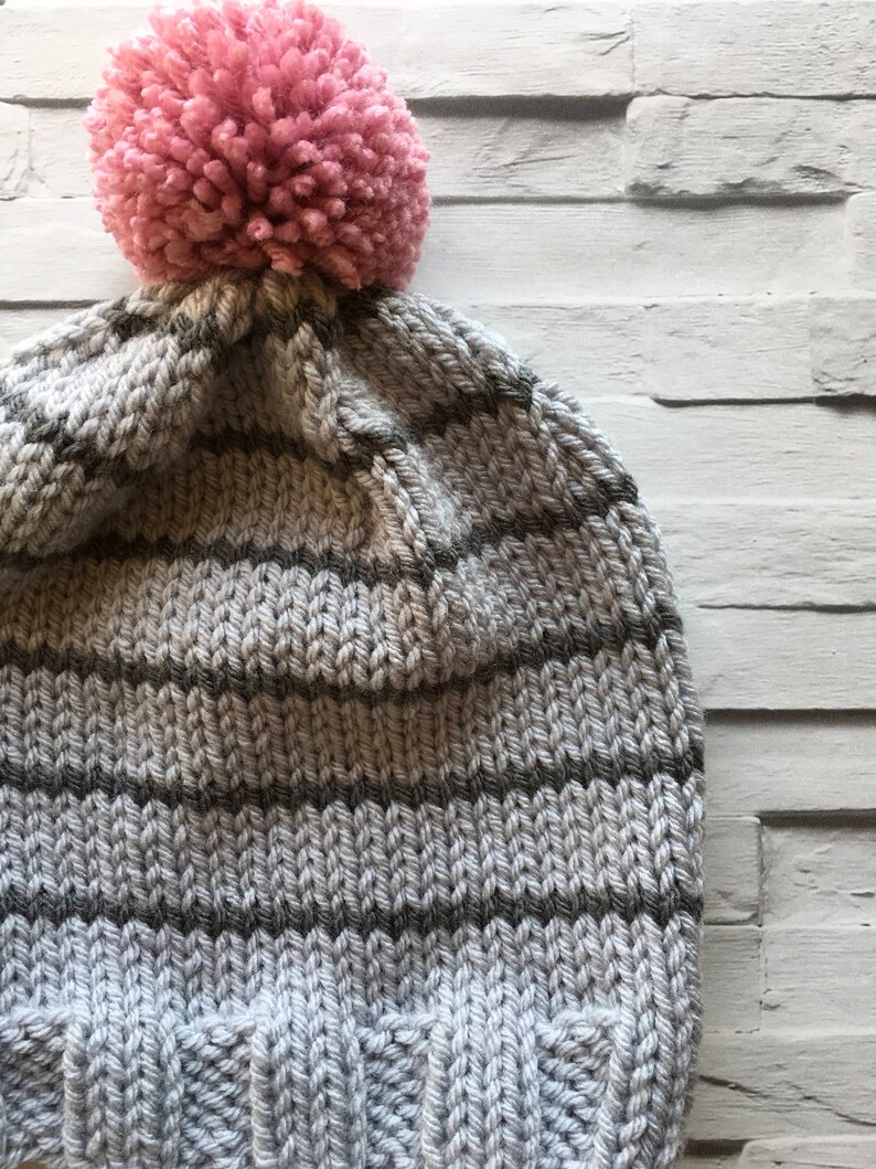 Puede incluir: Un gorro de punto con un borde acanalado y rayas horizontales en tonos alternos de gris claro y gris oscuro. La parte superior del gorro est&aacute; rematada con un pomp&oacute;n grande, redondo y texturizado en un color rosa suave, creando un contraste definido.
