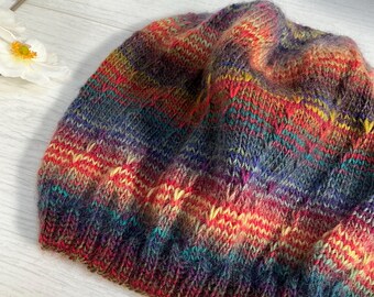 Könnte beinhalten: Handgestrickte Beanie-Mütze in einer lebendigen Farbmischung aus Rot, Gelb, Blau und Lila. Die Mütze hat ein strukturiertes Strickmuster und ein weiches, gemütliches Aussehen, ideal für kaltes Wetter.