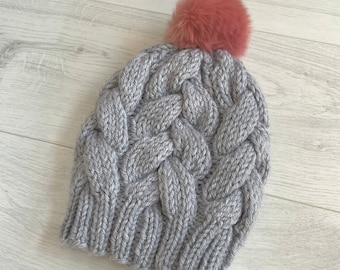 Könnte beinhalten: Graue Zopfmuster-Beanie-Mütze mit einem rosa Kunstfell-Bommel. Die Mütze hat ein dickes, strukturiertes Zopfmuster und ist aus weichem, warmem Garn gefertigt. Ein stilvolles Winteraccessoire.