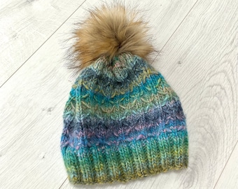 Könnte beinhalten: Eine handgestrickte Beanie-Mütze mit einem Kunstfell-Bommel. Die Mütze hat ein strukturiertes Strickmuster und ist mit Bändern in Blau, Grün, Lila und Gelb gefärbt. Der Bommel ist hellbraun.