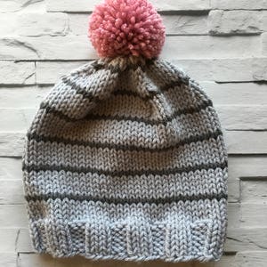 Puede incluir: Un gorro tejido a mano con un borde acanalado y un cuerpo con rayas horizontales alternas en gris claro y gris oscuro. La parte superior del gorro est&aacute; rematada con un pomp&oacute;n grande, redondo y texturizado en un suave color rosa empolvado.