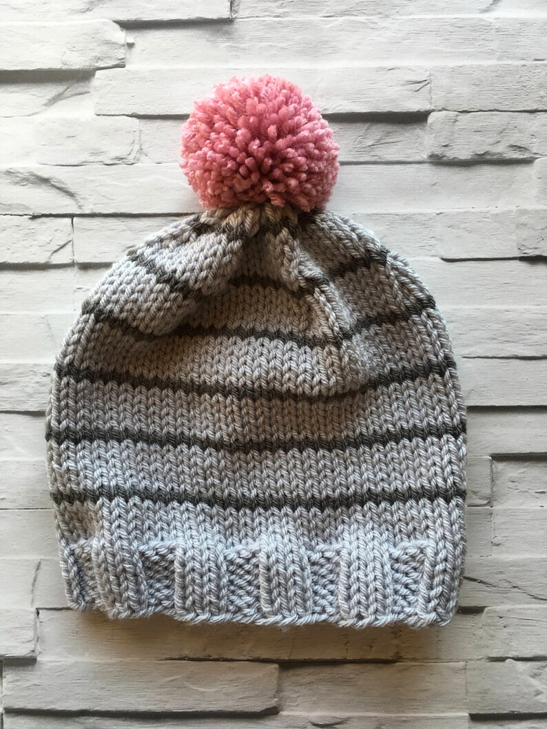 Puede incluir: Un gorro tejido a mano con un borde acanalado y un cuerpo con rayas horizontales alternas en gris claro y gris oscuro. La parte superior del gorro est&aacute; rematada con un pomp&oacute;n grande, redondo y texturizado en un suave color rosa empolvado.