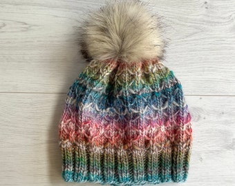 Könnte beinhalten: Eine handgestrickte Beanie-Mütze mit einem Kunstfell-Bommel. Die Mütze hat ein farbenfrohes, strukturiertes Muster mit Blau-, Rosa-, Grün- und Weißtönen. Der Bommel ist eine Mischung aus hellbraun und grau.