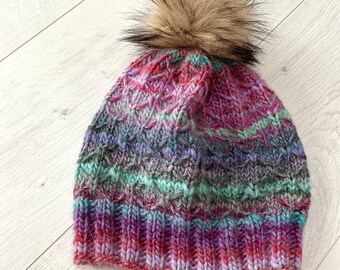 Könnte beinhalten: Eine handgestrickte Mütze in Rot-, Lila-, Grün- und Grautönen. Die Mütze hat ein strukturiertes Strickmuster und ist mit einem flauschigen, braunen Kunstfell-Bommel versehen. Dieses Winteraccessoire ist gemütlich.