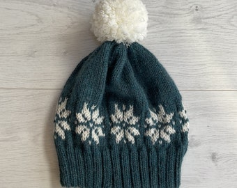 Könnte beinhalten: Ein tealfarbener Strickhut mit weißen Schneeflockenmustern und einem flauschigen weißen Bommel oben. Der Hut ist aus dickem, strukturiertem Garn gefertigt. Das Schneeflocken-Design verleiht diesem Winteraccessoire eine festliche Note.