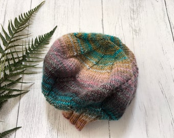 Puede incluir: Un gorro de punto con un patrón de degradado de colores en tonos rosa, turquesa, marrón y amarillo. El gorro está hecho con un hilo suave y tiene un ajuste holgado.