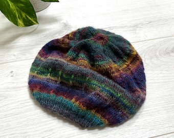 Könnte beinhalten: Eine handgestrickte Beanie-Mütze mit einem farbenfrohen Streifenmuster. Die Mütze hat eine Mischung aus Farben, darunter Blau, Lila, Grün und Gelb. Die Mütze ist aus weichem, strukturiertem Garn gefertigt.