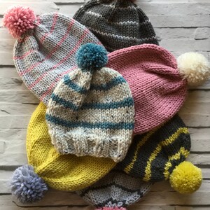 Puede incluir: Una colecci&oacute;n de siete gorros de punto en varios colores, incluyendo gris, amarillo, rosa y crema. Cada gorro presenta un borde acanalado y un pomp&oacute;n de lana en la parte superior. Los dise&ntilde;os incluyen colores s&oacute;lidos, rayas y patrones moteados.