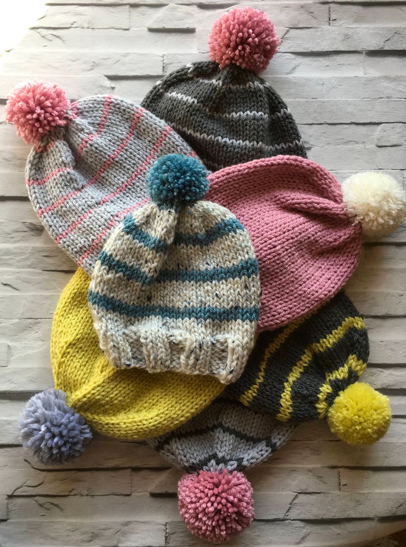 Puede incluir: Una colecci&oacute;n de siete gorros de punto en varios colores, incluyendo gris, amarillo, rosa y crema. Cada gorro presenta un borde acanalado y un pomp&oacute;n de lana en la parte superior. Los dise&ntilde;os incluyen colores s&oacute;lidos, rayas y patrones moteados.