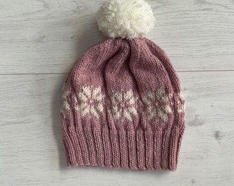 Könnte beinhalten: Eine gestrickte Wintermütze in Staubrosa mit weißen Schneeflockenmustern. Die Mütze hat einen gerippten Bund und einen großen weißen Bommel oben. Die Mütze ist aus weichem Garn gefertigt, ideal für kaltes Wetter.