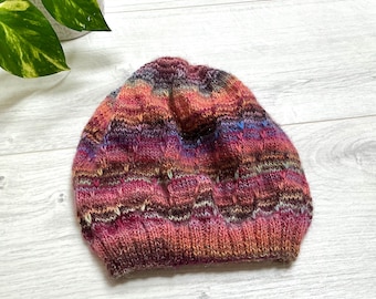 Könnte beinhalten: Handgestrickte Beanie-Mütze in Rosa, Lila, Orange und Braun. Die Mütze hat einen gerippten Bund und eine strukturierte Krone. Das Garn ist wahrscheinlich eine Wollmischung. Durchmesser ca. 20 cm.