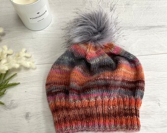 Könnte beinhalten: Ein mehrfarbiger Strickmütze mit einem großen grauen Kunstfell-Bommel. Die Mütze hat Streifen in Orange, Rosa, Grau und Braun. Diese warme Mütze ist ein Winteraccessoire, perfekt für kalte Tage.