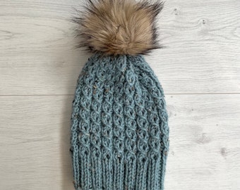 Könnte beinhalten: Blauer Strick-Beanie mit strukturiertem Muster und einem großen, flauschigen, braunen Kunstfell-Bommel. Die Mütze ist aus gesprenkeltem Garn gefertigt und für kaltes Wetter konzipiert, ca. 20 cm hoch.